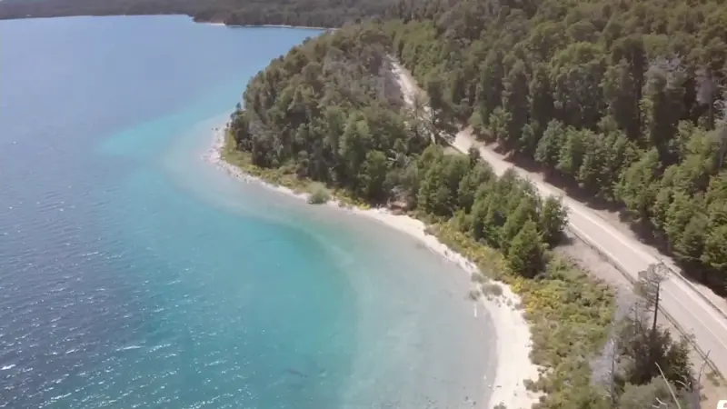 La costa del lago Nahuel Huapi, de agua turquesa, bordeada por el bosque y un camino, vista desde el aire