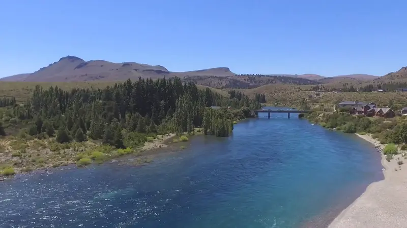 Un río de agua cristalina entre bosques y estepa, con un puente al fondo